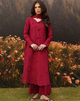 A woman stands outdoors wearing a long-sleeved, high-neck maroon kurta with matching pants. The kurta features a high mock neck with a white inner collar, button details down the front, and floral embroidery near the hem.