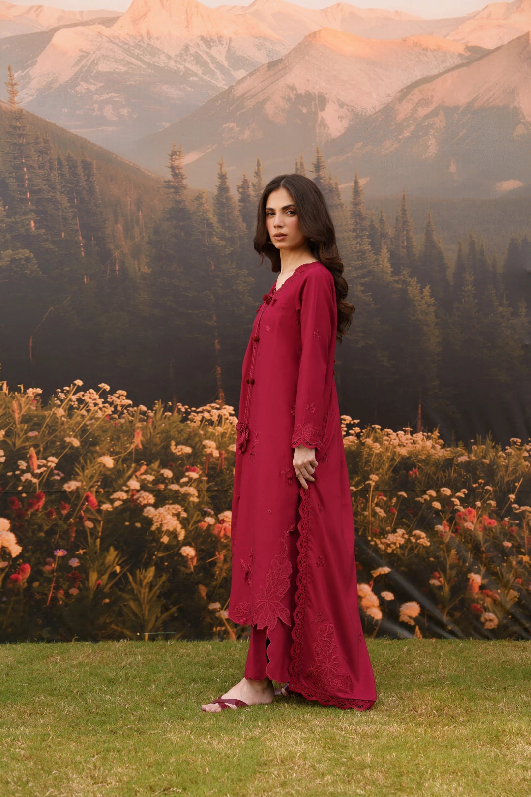 A woman stands outdoors in a mountain landscape wearing a long, deep magenta-red dress. The background features a grassy lawn, a dense bed of pink and white flowers, pine trees, and mountains under a warm sky.