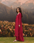 A woman stands outdoors in a mountain landscape wearing a long, deep magenta-red dress. The background features a grassy lawn, a dense bed of pink and white flowers, pine trees, and mountains under a warm sky.