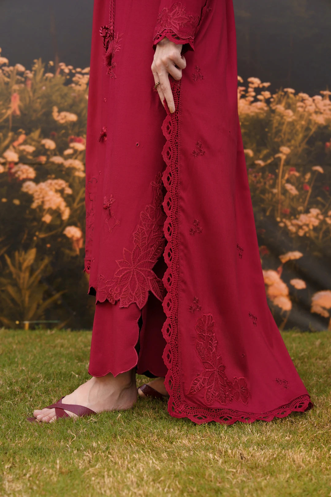 A person outdoors wearing a deep red embroidered kurta and matching pants, standing on grass with flowers blurred in the background. The kurta has lace trim and floral embroidery.