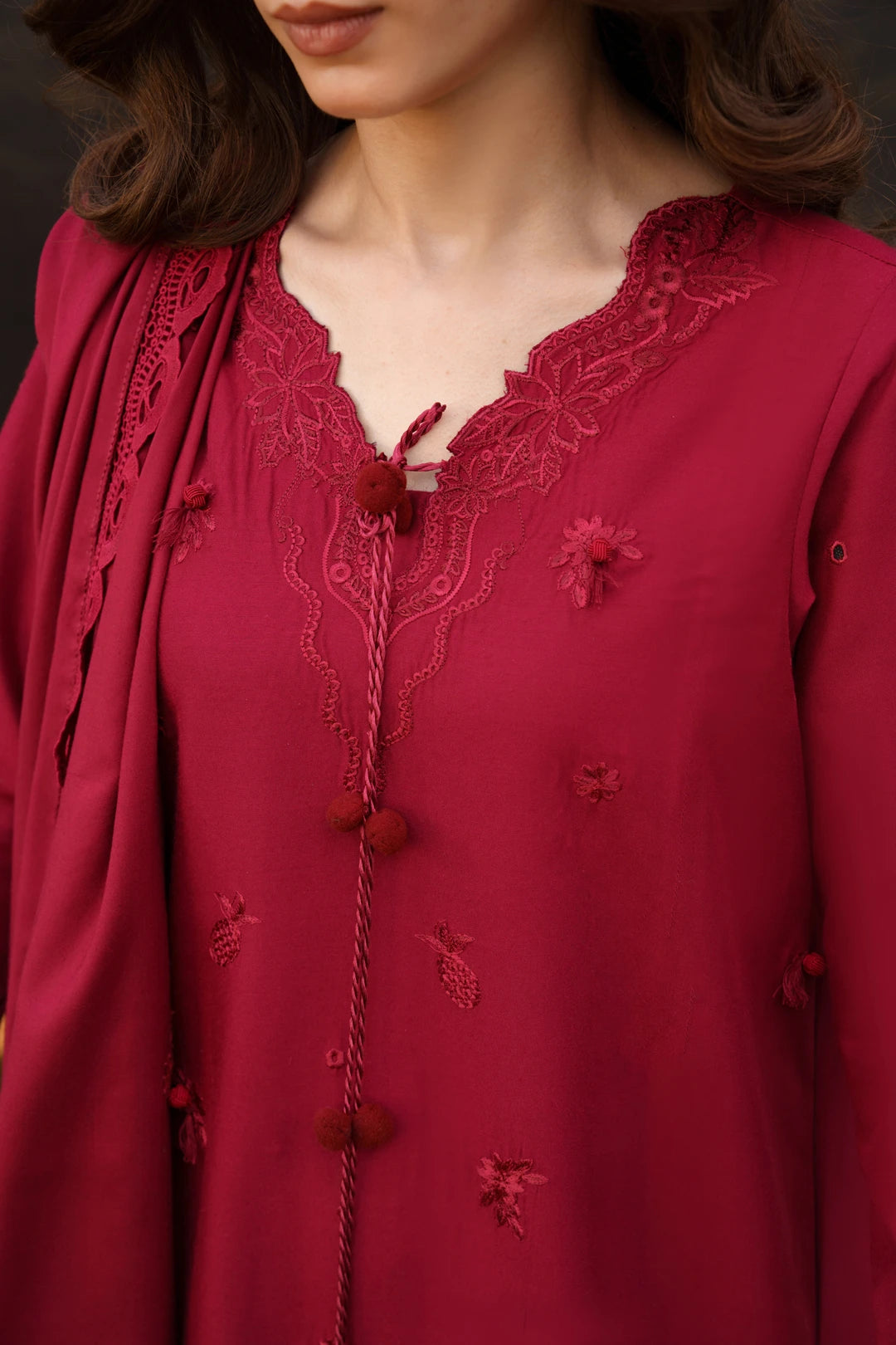 A close-up of a woman wearing a deep red embroidered tunic with lace trim, a matching draped scarf, and decorative pom-pom ties.