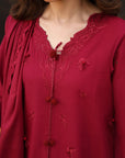 A close-up of a woman wearing a deep red embroidered tunic with lace trim, a matching draped scarf, and decorative pom-pom ties.