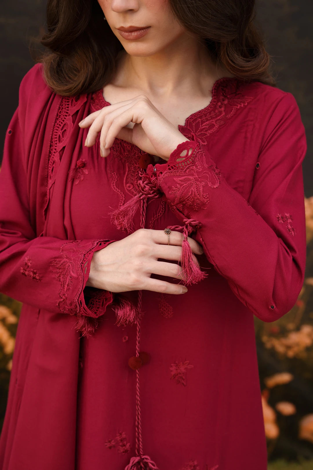 A woman wearing a long-sleeved deep red dress with intricate lace trim, tassel ties, and embroidered floral details, shown in a close-up shot against a blurred outdoor background.