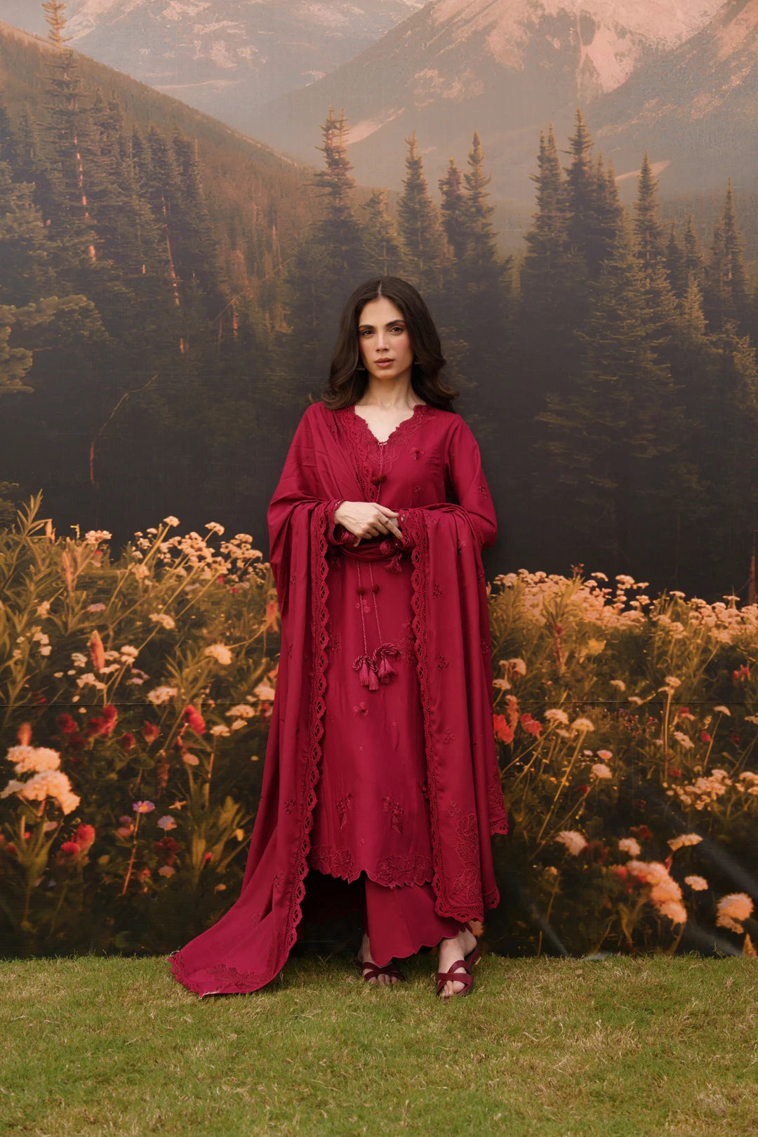 A woman stands outdoors in front of a scenic backdrop of mountains, pine trees, and flowers, wearing a long burgundy dress with wide draped sleeves and a V-neckline.