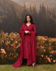 A woman stands outdoors in front of a scenic backdrop of mountains, pine trees, and flowers, wearing a long burgundy dress with wide draped sleeves and a V-neckline.