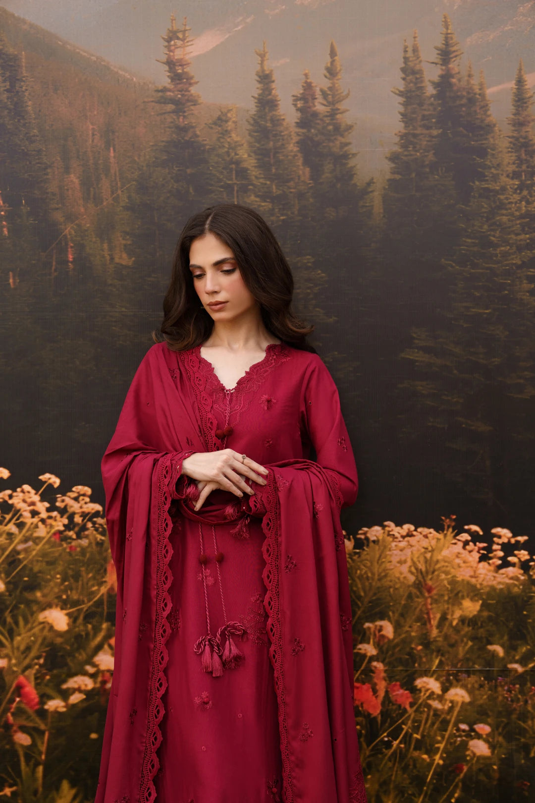 A woman wearing a long burgundy red dress with a matching draped shawl stands in a field of wildflowers with a forest and mountains in the background.
