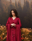 A woman wearing a long burgundy red dress with a matching draped shawl stands in a field of wildflowers with a forest and mountains in the background.