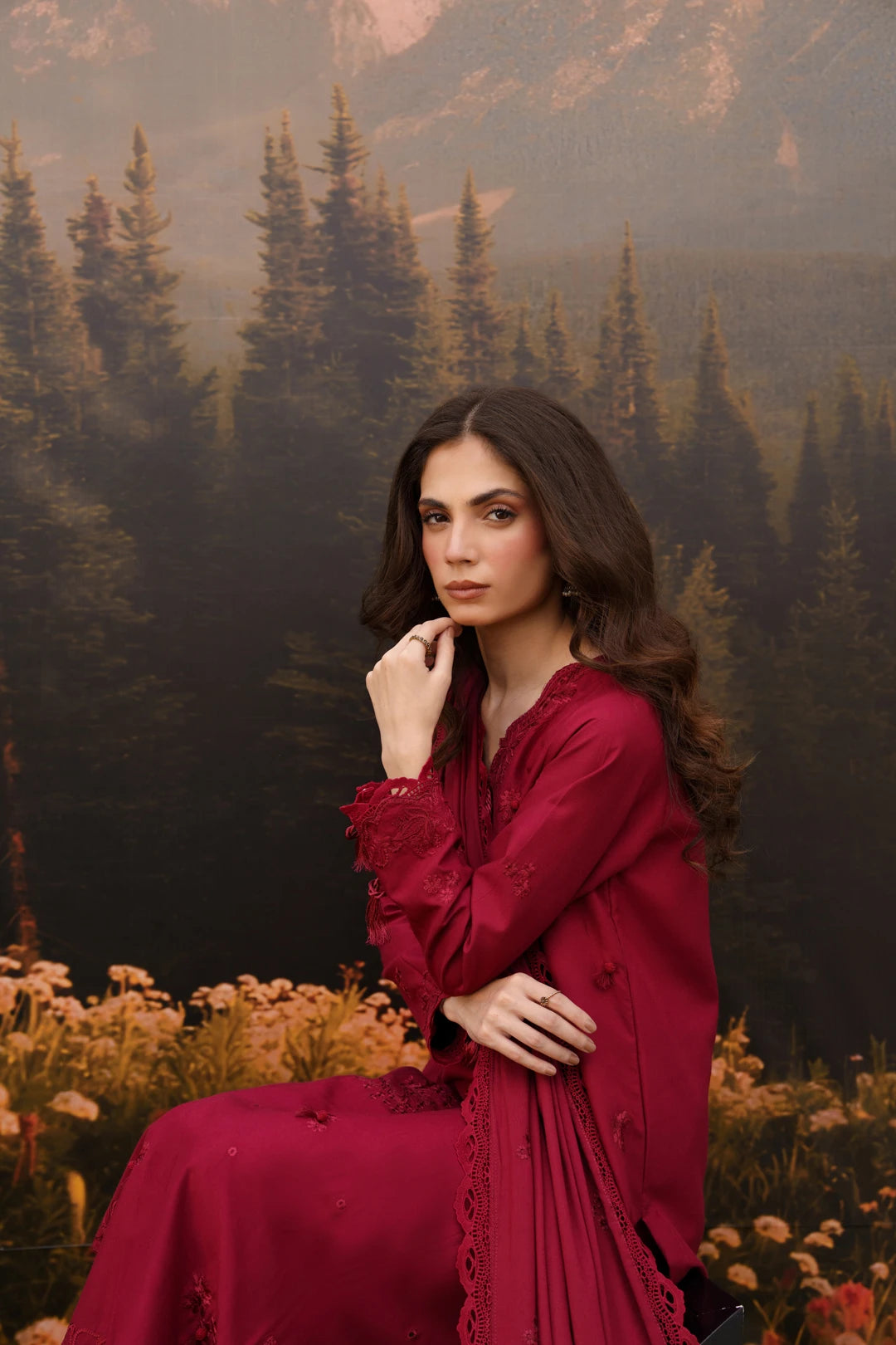 A woman wearing a deep burgundy red embroidered dress sits in front of a forest and mountain backdrop. The dress has long sleeves with lace detailing and a flowing skirt.