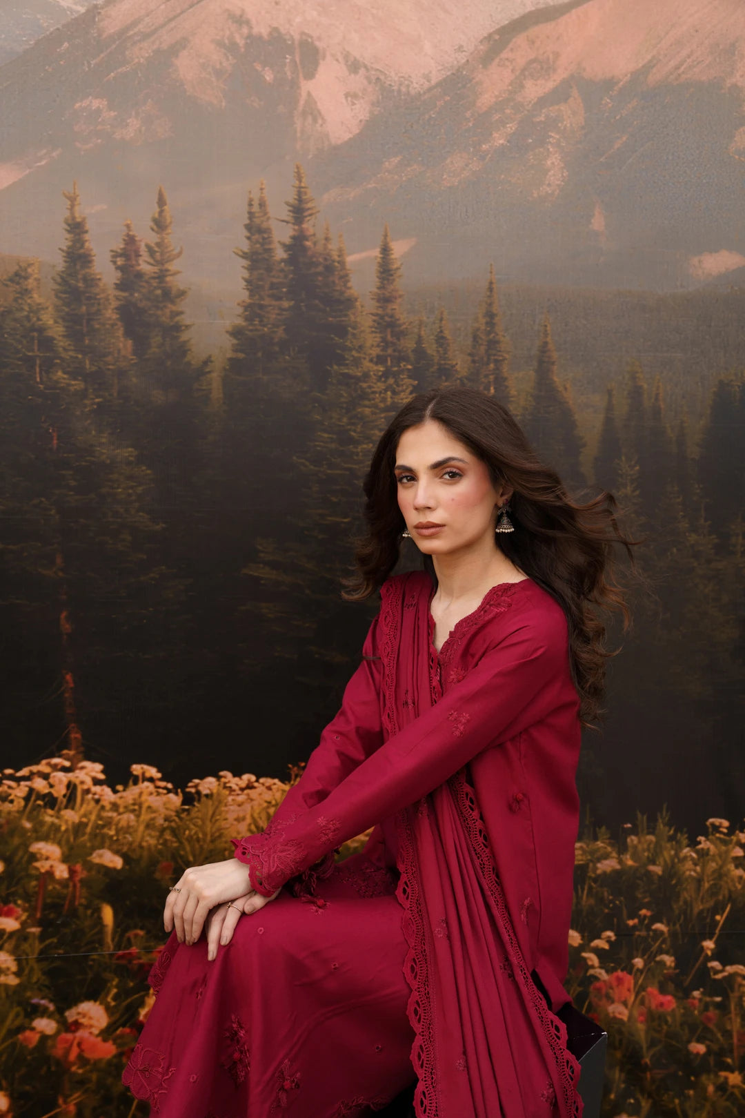 A woman wearing a deep magenta-red embroidered kurta sits in a field of wildflowers with a large scenic backdrop of a pine forest and mountains.