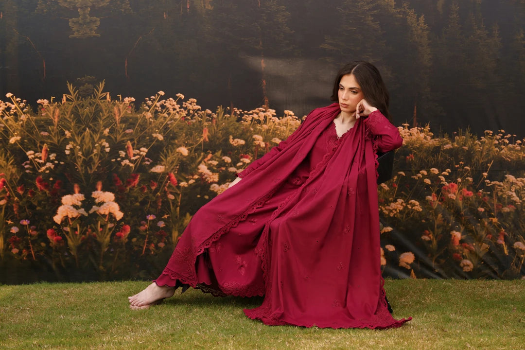 A woman wearing a flowing deep burgundy robe or nightgown sits barefoot on grass in front of a floral backdrop. The garment has a loose, draped silhouette with long sleeves and a deep V-neckline.