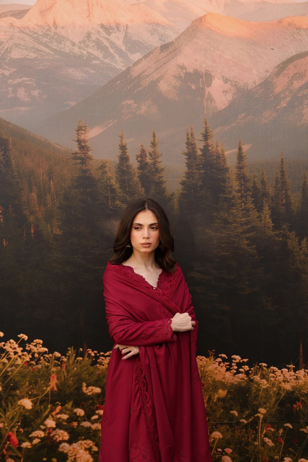 A woman stands in a mountain meadow wearing a long burgundy wrap dress, with a large scenic backdrop of pine trees and snow-capped mountains behind her.
