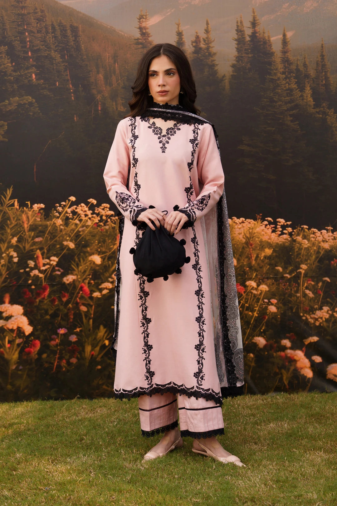 A woman stands outdoors on grass in front of a floral backdrop and forested mountains, wearing a light pink traditional outfit with black embroidered detailing and a black dupatta. She holds a small black pouch.