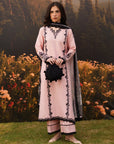 A woman stands outdoors on grass in front of a floral backdrop and forested mountains, wearing a light pink traditional outfit with black embroidered detailing and a black dupatta. She holds a small black pouch.