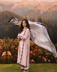 A woman stands outdoors in front of mountains and a flower bed, wearing a long light pink embroidered kurta with a matching sheer scarf flowing behind her.