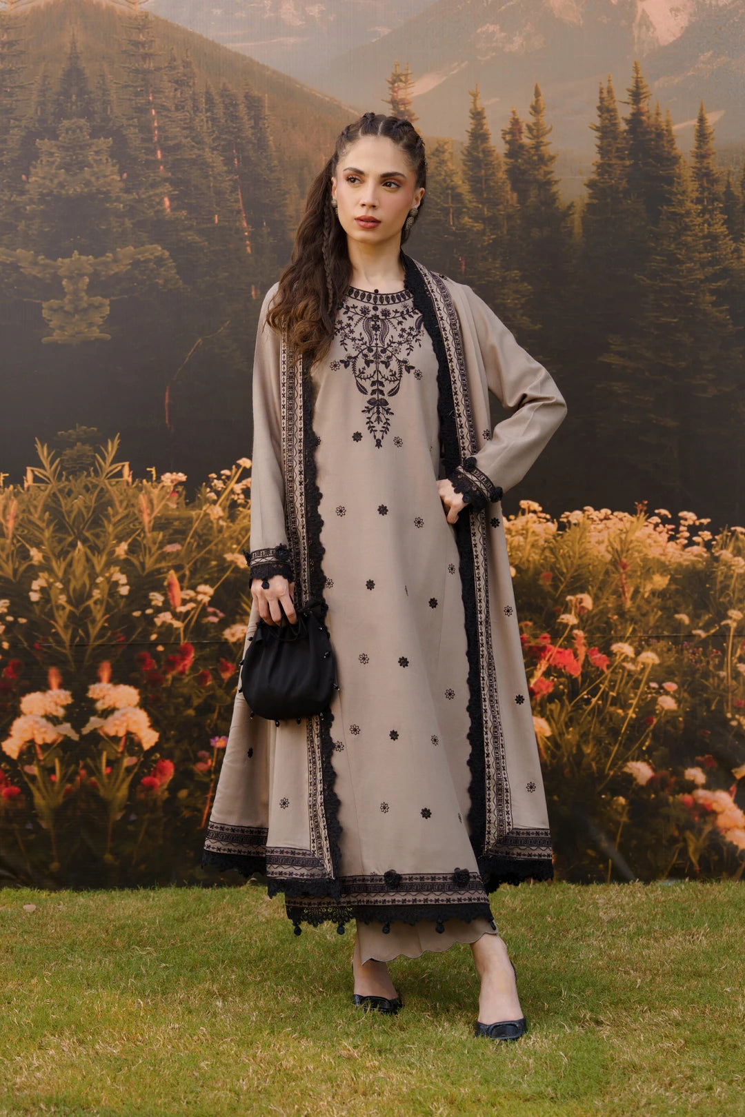 A woman stands outdoors in front of a scenic mountain and forest backdrop, wearing a long grey traditional outfit with black embroidery, black polka dots, and a matching black outer layer. She is holding a small black handbag.
