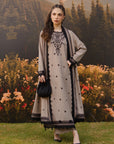 A woman stands outdoors in front of a scenic mountain and forest backdrop, wearing a long grey traditional outfit with black embroidery, black polka dots, and a matching black outer layer. She is holding a small black handbag.