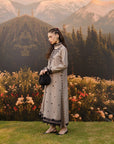 A woman stands outdoors in front of mountains and a flowered backdrop, wearing a long grey embroidered dress with black trim and holding a small black handbag.