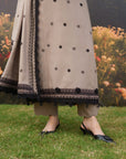A person outdoors wearing a grey traditional outfit with black embroidered floral motifs, a black-trimmed border, and black slingback heels, standing on grass with flowers blurred in the background.