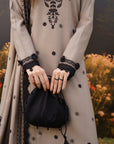 A person wearing a grey embroidered traditional outfit with black floral detailing and lace cuffs, holding a small black drawstring pouch bag, with a blurred outdoor garden background.