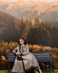 A woman wearing a traditional long kurta set in taupe with black embroidery and a matching trouser sits on a dark wooden bench in front of a scenic mountain backdrop.