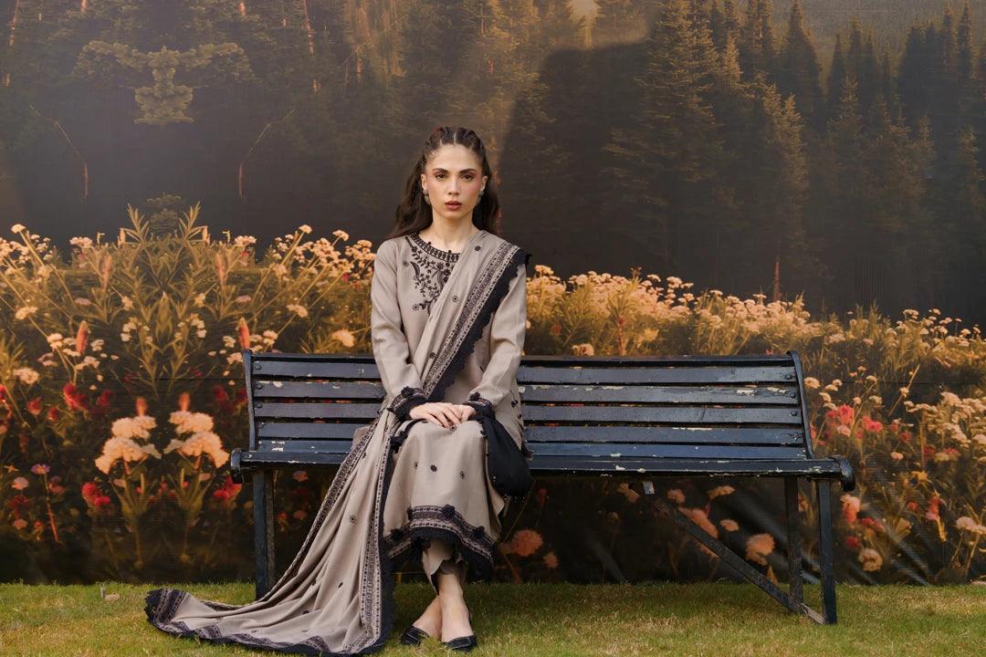 A woman sits on a dark wooden bench in front of a floral and forest backdrop, wearing a traditional outfit with a light gray kurta and a matching draped scarf with black embroidery.