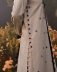 A close-up of a woman wearing a long grey tunic with black floral embroidery, black buttons down the front, and a black embroidered border, set against a blurred garden background.