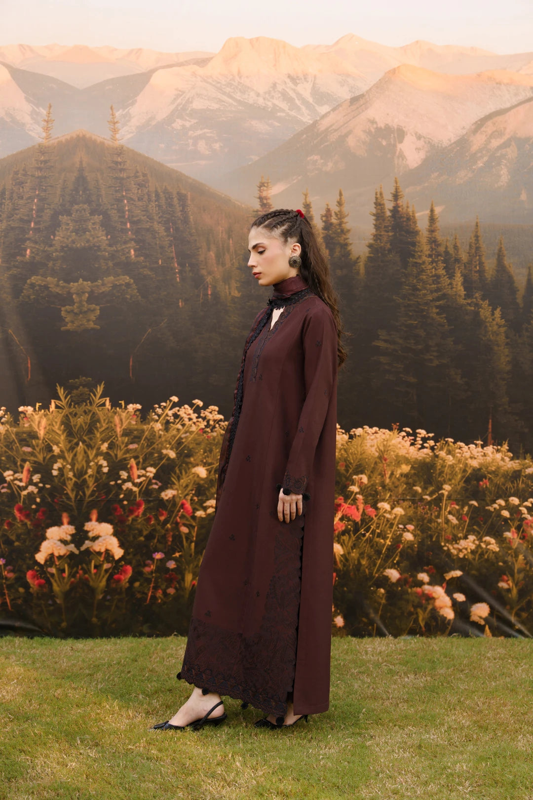 A woman stands in profile outdoors in front of a mountain-and-flower backdrop, wearing a long dark burgundy maxi dress with a high neck and side slit.