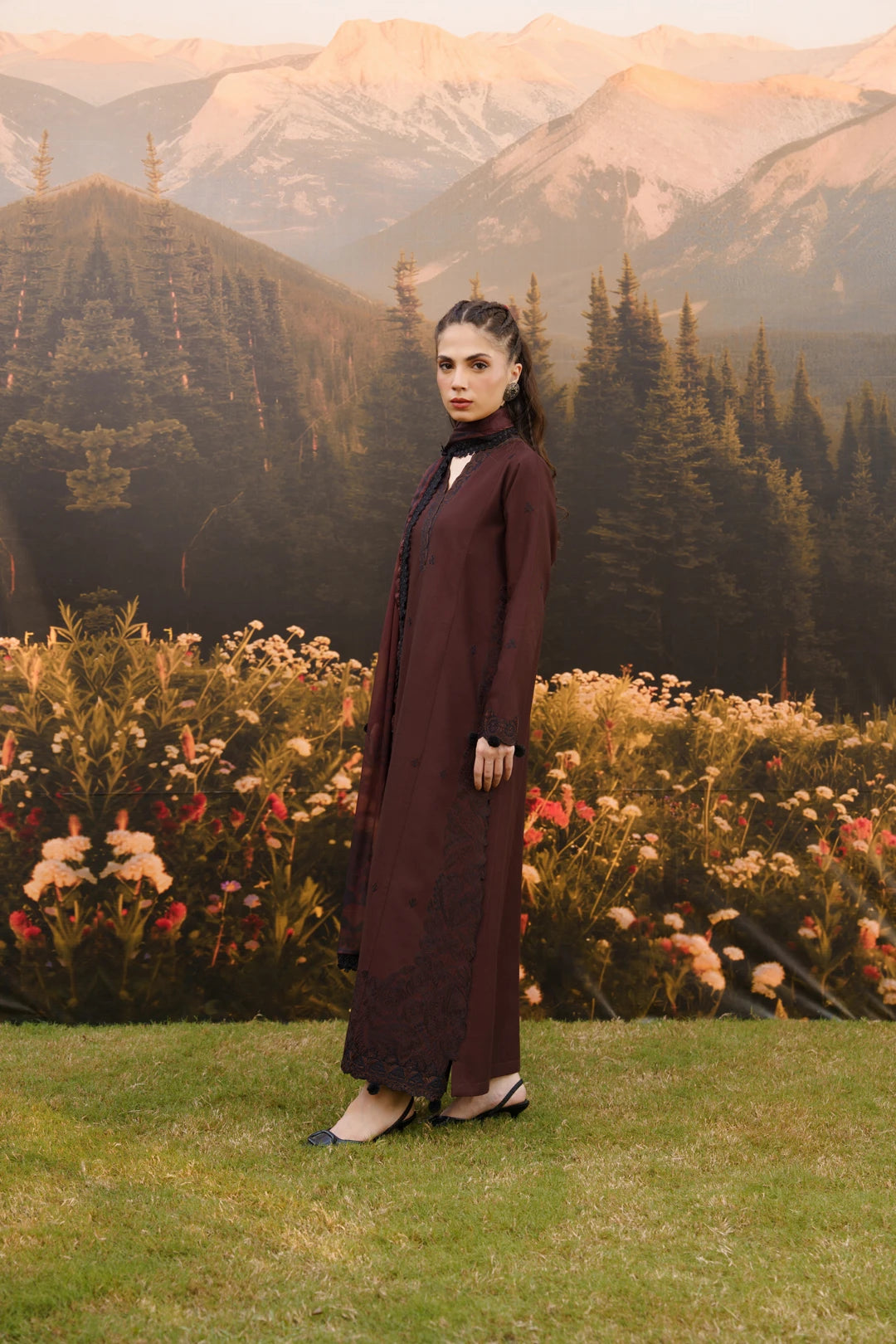 A woman stands outdoors in front of mountains, trees, and flowers, wearing a long dark burgundy dress with a high neck and sheer embroidered lower hem.