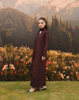 A woman stands outdoors in front of mountains, trees, and flowers, wearing a long dark burgundy dress with a high neck and sheer embroidered lower hem.