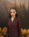 A woman wearing a dark plum-colored traditional tunic with black lace trim at the V-neck, standing in front of a scenic backdrop of pine trees and mountains.