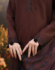 A close-up of a person wearing a dark brown traditional outfit with black embroidered details, lace trim, and pom-pom cuffs, holding a matching patterned dupatta against a blurred outdoor background.