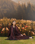 A woman sits cross-legged on a grassy field in front of a backdrop of wildflowers, pine trees, and mountains. She is wearing a long-sleeved dark plum purple robe with a matching draped scarf.