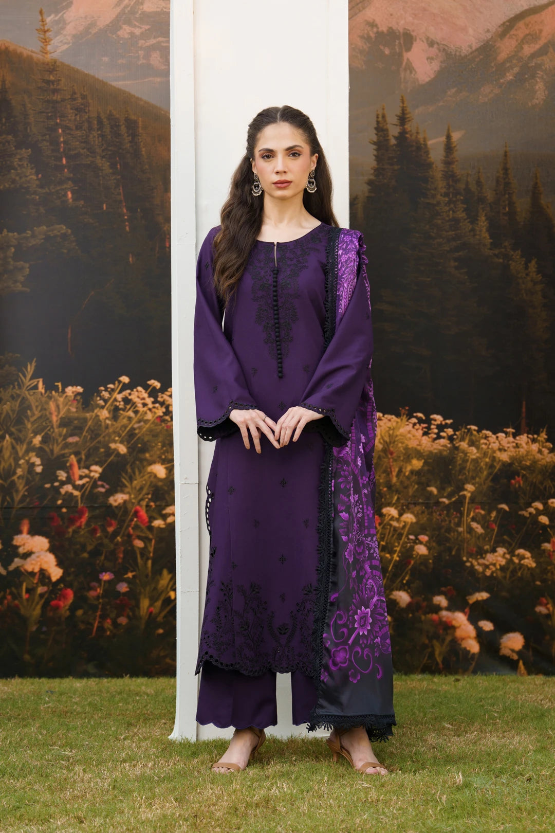 A woman stands outdoors wearing a deep purple traditional outfit with black embroidered details and a matching sheer purple dupatta. The background features a scenic mural of mountains, trees, and flowers, with a white vertical panel behind her.