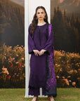 A woman stands outdoors wearing a deep purple traditional outfit with black embroidered details and a matching sheer purple dupatta. The background features a scenic mural of mountains, trees, and flowers, with a white vertical panel behind her.