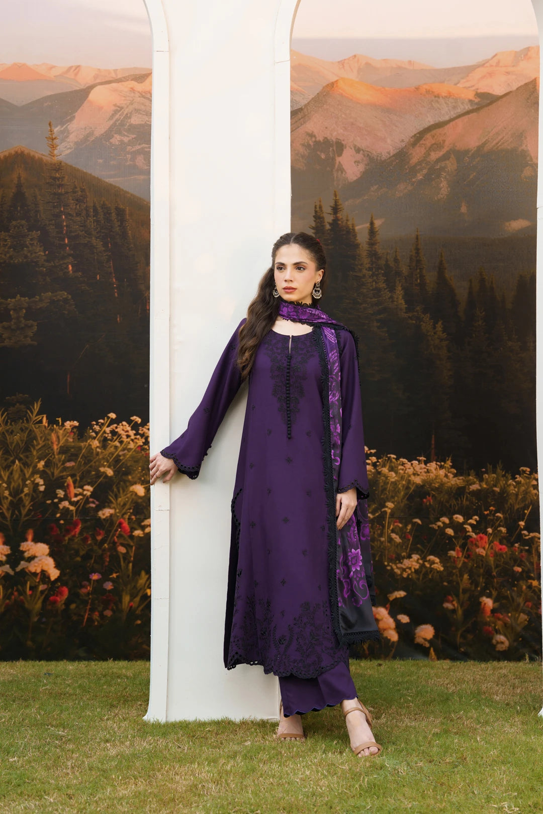A woman models a deep purple traditional South Asian outfit with matching pants and dupatta, standing next to a white column in front of a scenic backdrop of mountains and flowers.