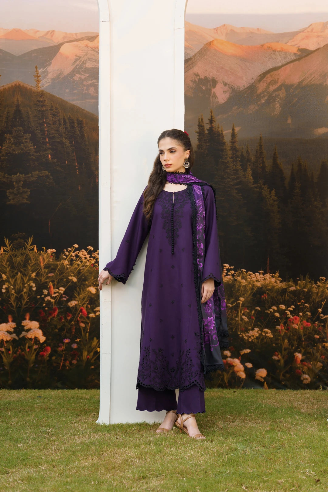 A woman modeling a deep purple traditional outfit with matching embroidered dupatta, standing next to a white column in front of a scenic mountain backdrop.