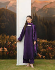 A woman modeling a deep purple traditional outfit with matching embroidered dupatta, standing next to a white column in front of a scenic mountain backdrop.