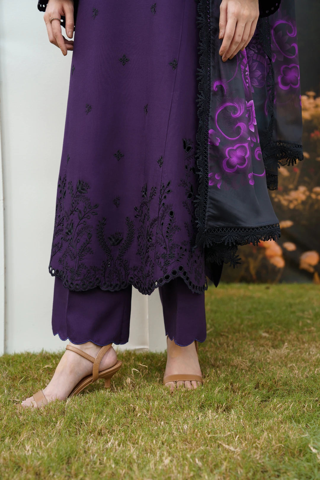 A person outdoors wearing a purple traditional outfit with black embroidered detailing, matching purple pants, and a sheer black dupatta with purple floral embroidery, standing on grass.