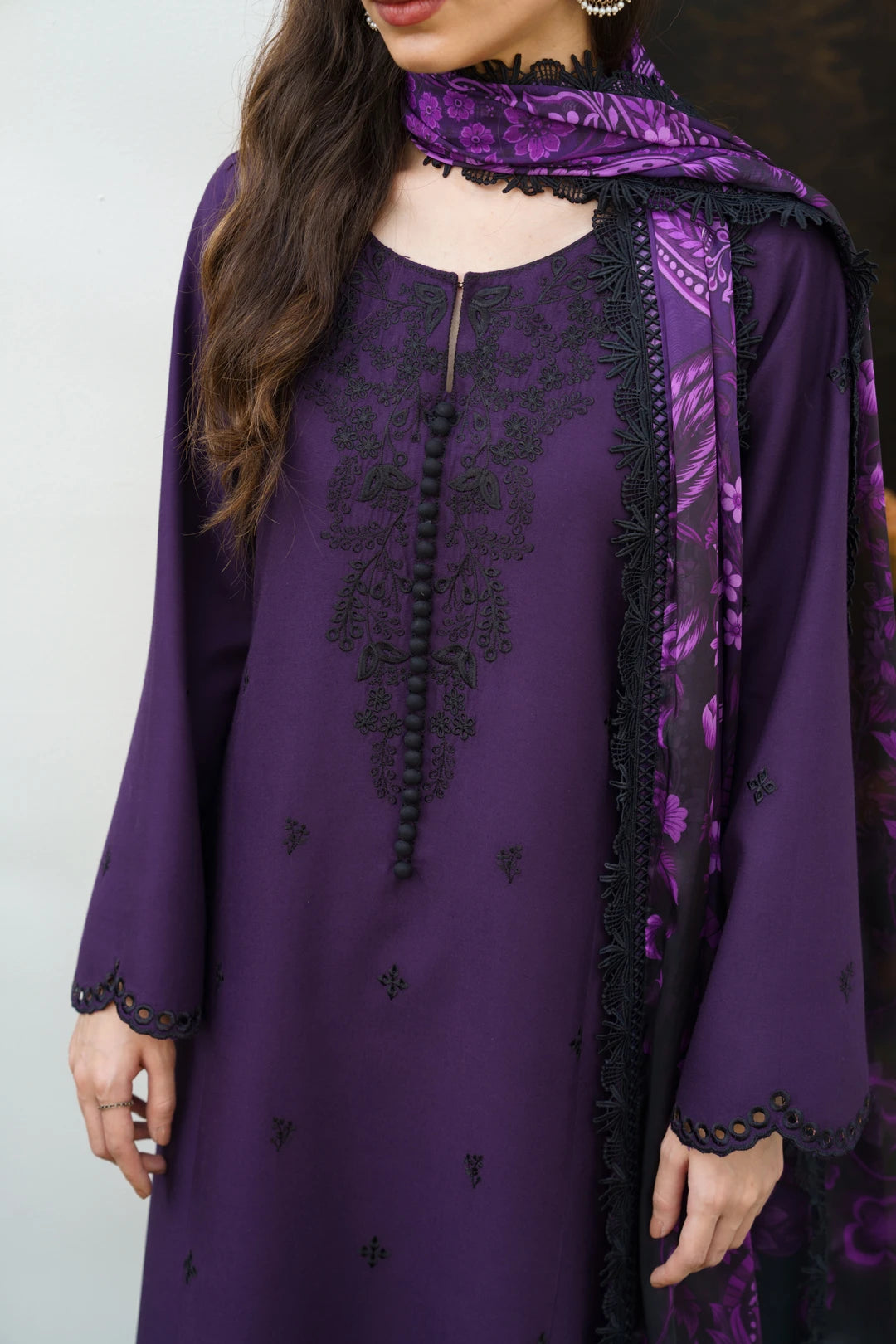 A woman wearing a purple traditional outfit with black embroidered details, a black tassel tie, and a matching purple patterned dupatta.
