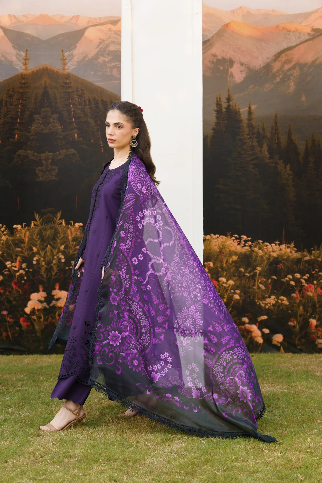 A woman models a traditional South Asian outfit consisting of a deep purple kurta and matching trousers, paired with a sheer black dupatta featuring intricate purple floral embroidery, set against a scenic backdrop of mountains and grass.