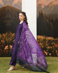 A woman models a traditional South Asian outfit consisting of a deep purple kurta and matching trousers, paired with a sheer black dupatta featuring intricate purple floral embroidery, set against a scenic backdrop of mountains and grass.