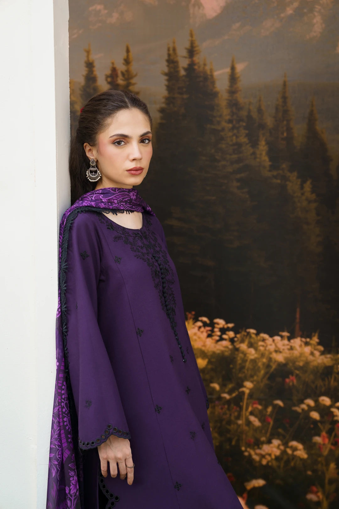 A woman models a deep purple traditional outfit with black embroidered details and a matching purple patterned dupatta, posed against a scenic forest mural background.