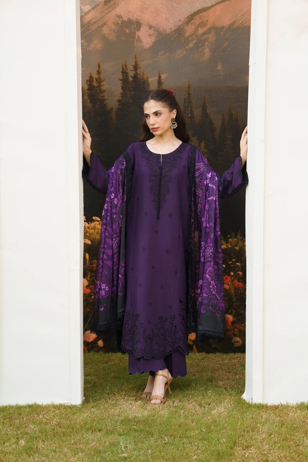 A woman models a deep purple traditional outfit with matching embroidered sheer sleeves, standing in a doorway with a scenic mountain backdrop.