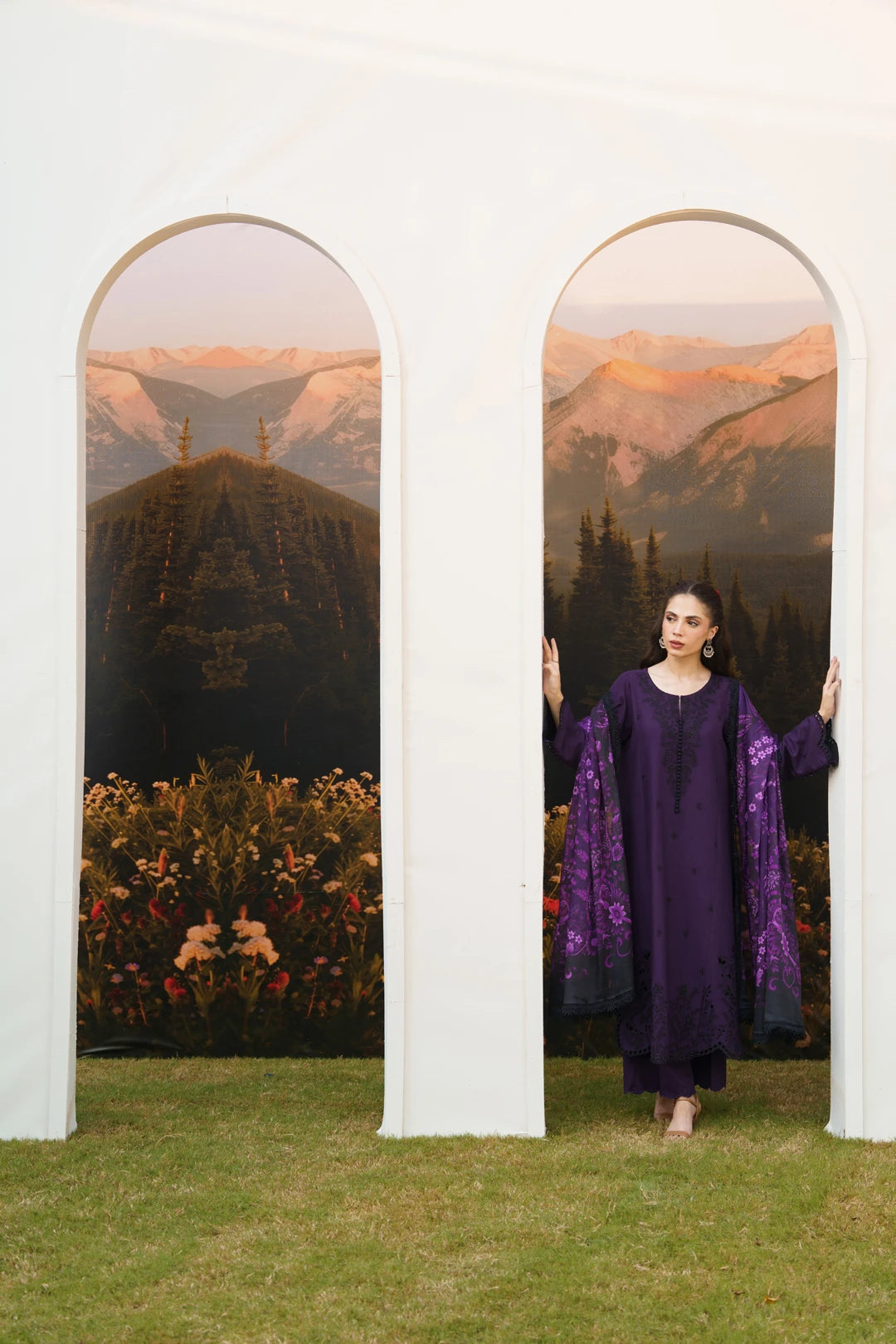 A woman stands between two large arched scenic panels, wearing a deep purple traditional outfit with patterned sleeves. The setting includes green grass at the bottom and a white architectural backdrop behind her.