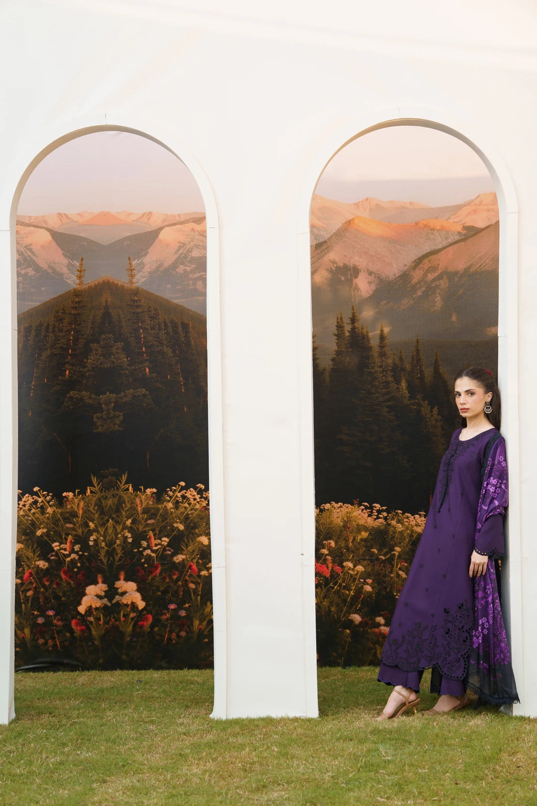 A woman wearing a deep purple embroidered kurta and matching trousers stands beside a white architectural backdrop with two arched cutouts showing a mountain landscape.