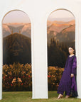 A woman wearing a deep purple embroidered kurta and matching trousers stands beside a white architectural backdrop with two arched cutouts showing a mountain landscape.