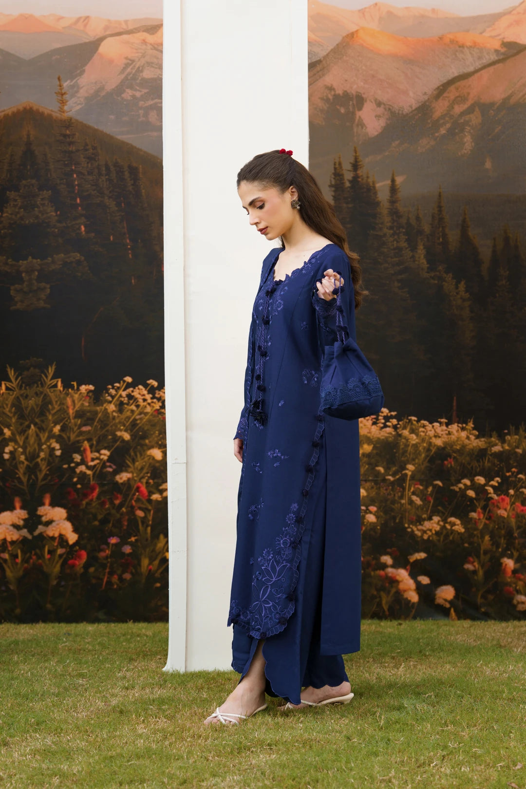 A woman models a deep navy blue embroidered kurta set with matching trousers and a matching handbag, standing outdoors in front of a scenic mountain backdrop.