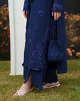 A person wearing a long navy blue embroidered tunic and matching pants, standing on grass and holding a matching navy drawstring bag.