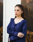 A woman wearing a dark navy blue long-sleeved dress with intricate floral embroidery, standing in front of a white pillar and a scenic backdrop.