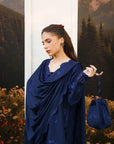 A woman wearing a dark navy blue embroidered shawl draped over her shoulders, holding a matching embroidered navy blue handbag, posed in front of a scenic backdrop of mountains and forests.